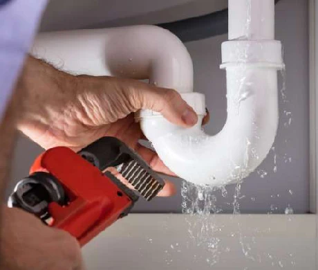 A water damage restoration specialist using a red wrench to fix a leaking white sink pipe, with water visibly dripping from the joint of the pipe.