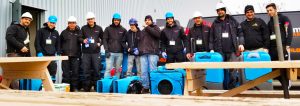A group of plumbers wearing helmets and work gear, posing happily with tools and equipment at a work site.