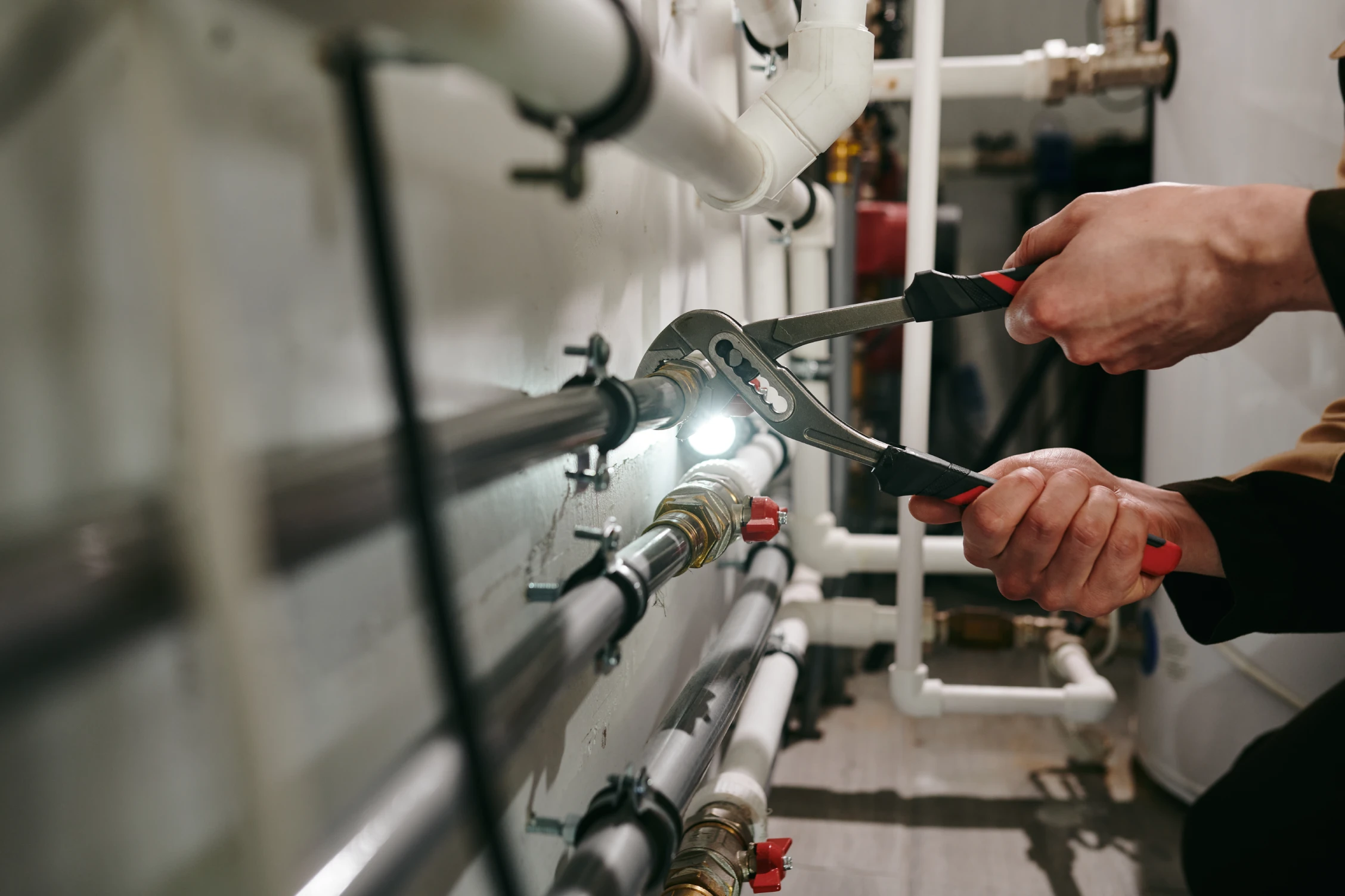 A plumber using a wrench to tighten a fitting on a pipe in a well-lit boiler room with several white pipes running along the wall.