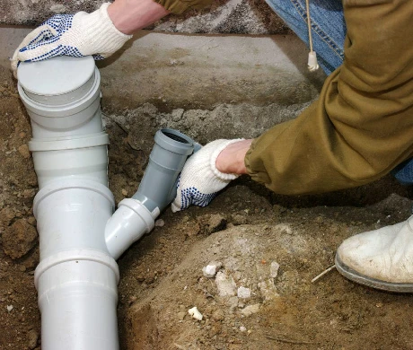 A plumber wearing gloves is assembling PVC plumbing pipes in a dirt trench, focusing on connecting a gray elbow joint to straight white pipes.