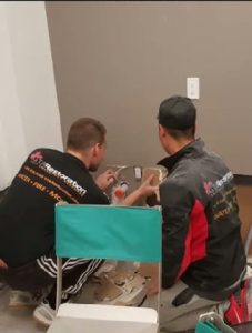 Two men in branded work shirts are installing or repairing an electrical outlet in a room with gray walls. Plumbing tools and materials are scattered on the floor around them.