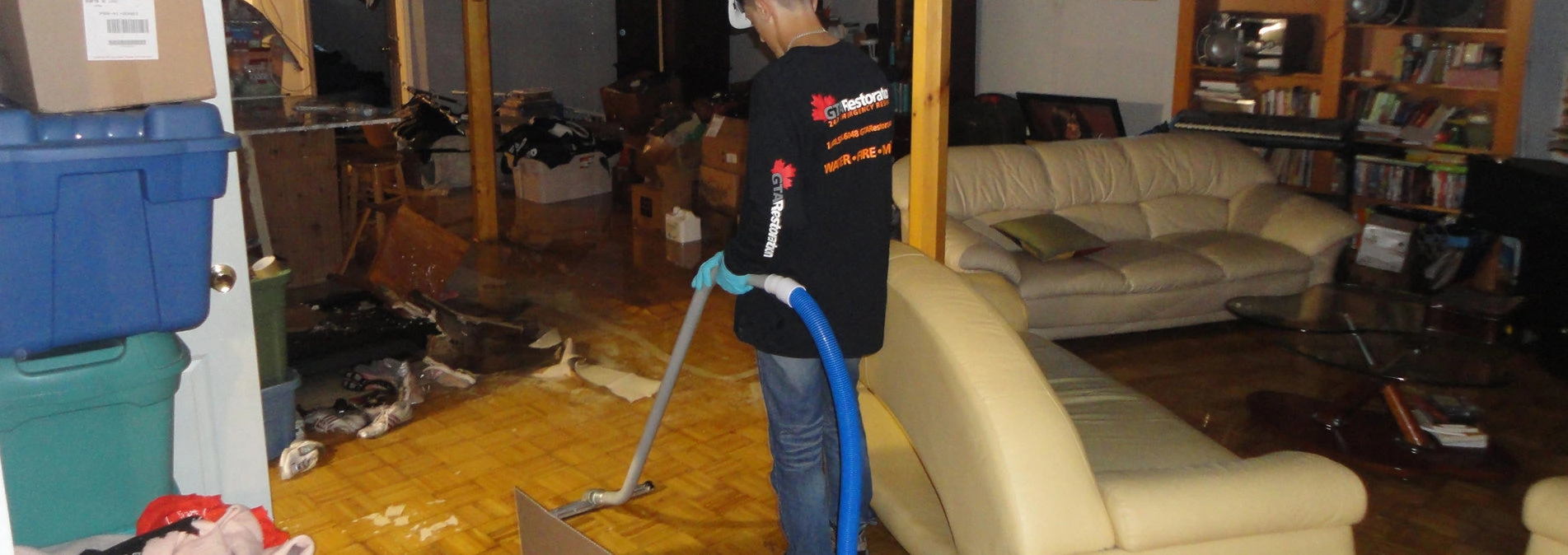 A person using a large vacuum hose during a water damage restoration in a cluttered living room with scattered items and furniture. They are wearing a black t-shirt with cleaning company logos.