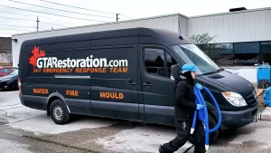 A plumber in a blue helmet carries a large hose past a black van branded with "gtarestoration.com" and logos indicating services for water damage restoration, fire, and mold damage. The scene is