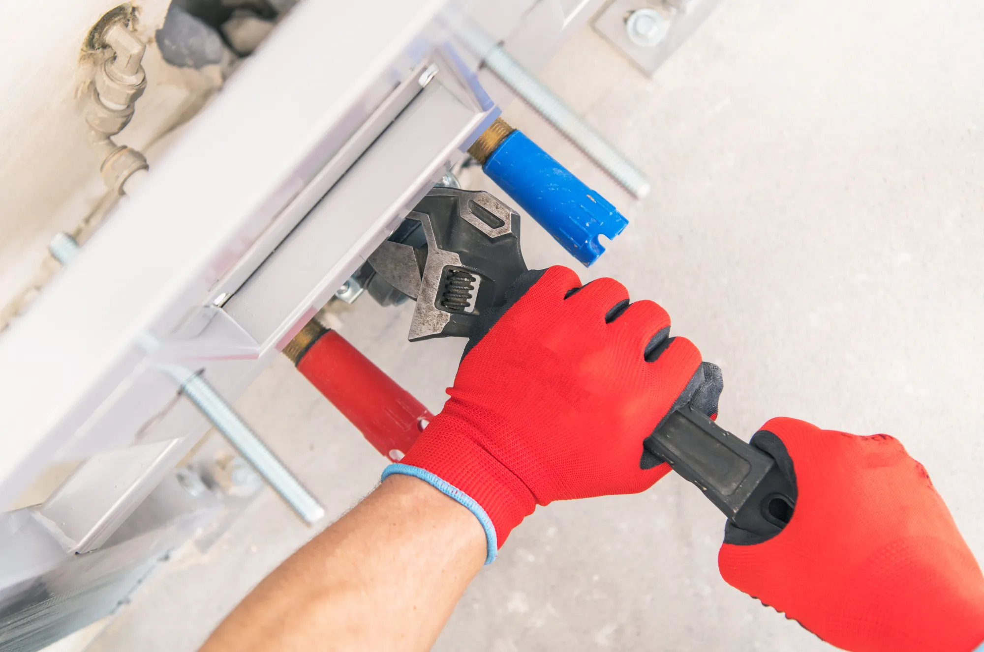 A plumber wearing red gloves uses a wrench to adjust or fix something underneath a white cabinet, with various tools visible.