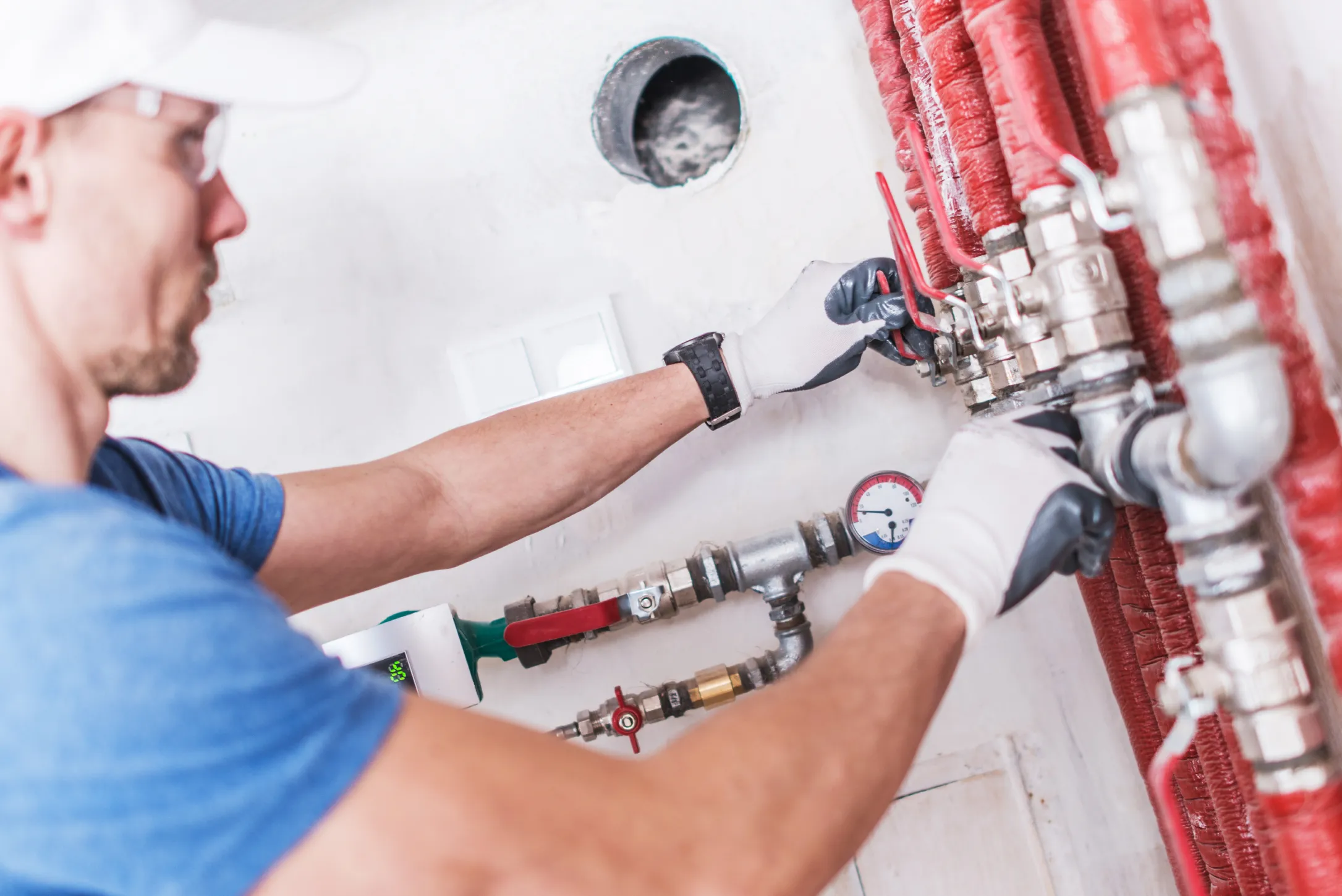 A plumber specializing in water damage restoration, wearing a blue t-shirt and white gloves, works on fixing red pipes and valves on a white wall, with tools in hand, focusing intently on his task