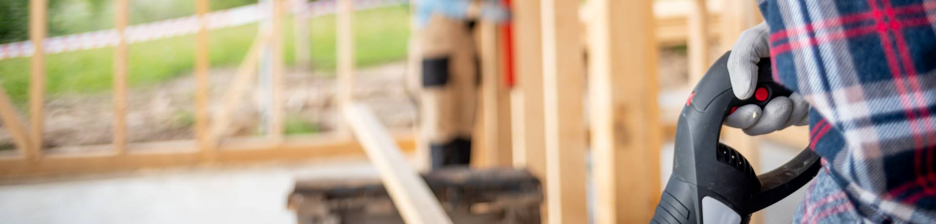 A close-up side view of a plumber wearing a plaid shirt holding a power tool handle at a construction site, with a partially built wooden structure in the blurry background.