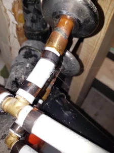 Close-up image showing a plumber working on a plumbing connection with copper and white PVC pipes, fitted with valves and connectors, located in a tight, dusty space.