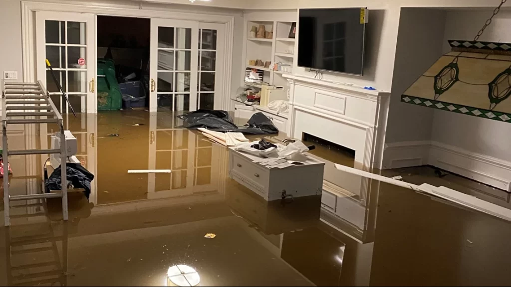 A flooded living room with water covering the floor requires urgent water damage restoration; furniture, books, and debris scattered around. Two white French doors and a built-in white cabinet are visible in the background.