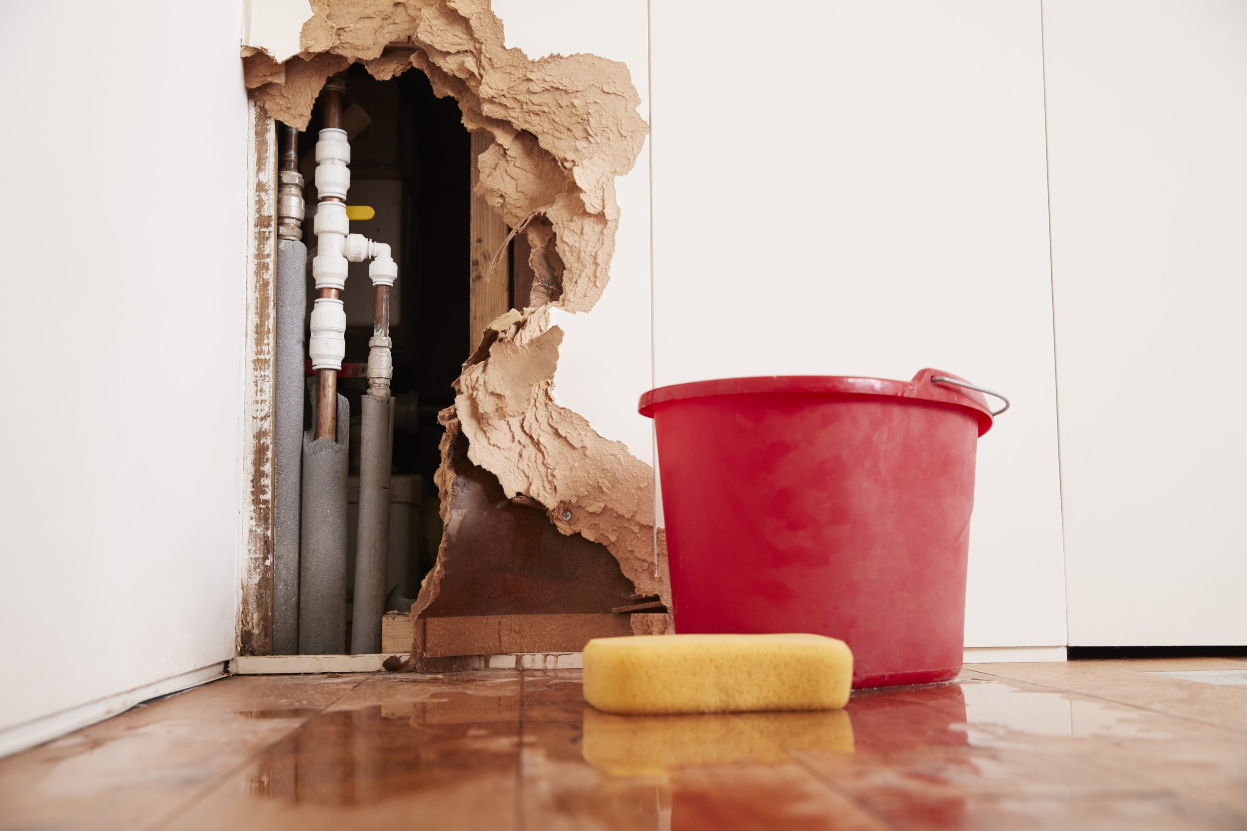 Red bucket and a yellow sponge on a tiled floor in front of a broken wall with visible pipes indicating water damage, suggesting the need for water damage restoration.