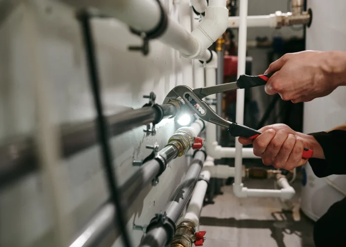 A plumber using a wrench to tighten a fitting on a pipe in a well-lit boiler room with several white pipes running along the wall.