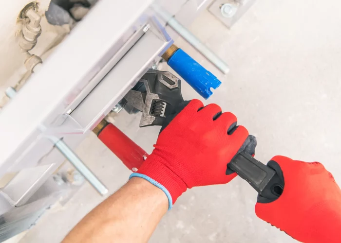 A plumber wearing red gloves uses a wrench to adjust or fix something underneath a white cabinet, with various tools visible.